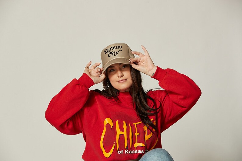 Pu&ograve; includere: Una donna indossa una felpa rossa con la scritta "CHIEF of Kansas" stampata e un cappello da camionista beige con "Kansas City" ricamato in nero.