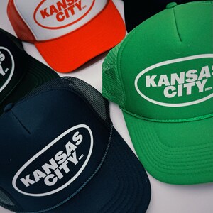 May include: Five trucker hats with a white oval logo that says "KANSAS CITY, MO". The hats are in different colours: red, black, green, blue, and dark green.
