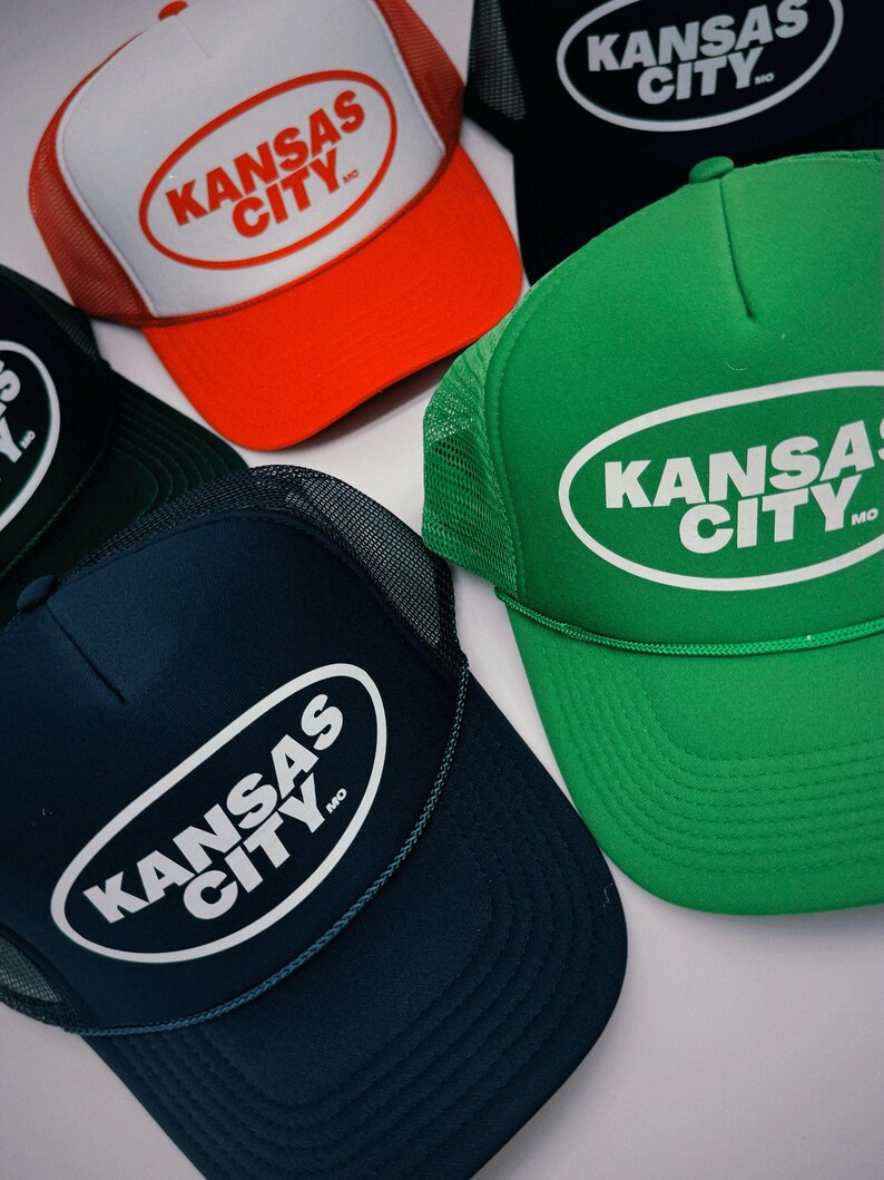 May include: Five trucker hats with a white oval logo that says "KANSAS CITY, MO". The hats are in different colours: red, black, green, blue, and dark green.