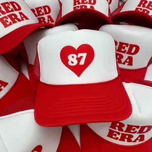 Puede incluir: Una pila de gorras de camionero rojas y blancas con un corazón y el número 87 en una gorra. Las otras gorras tienen las palabras "Red Era" en ellas.