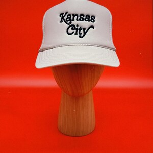 Pu&ograve; includere: Un cappello da camionista beige con un logo ricamato nero che dice "Kansas City".