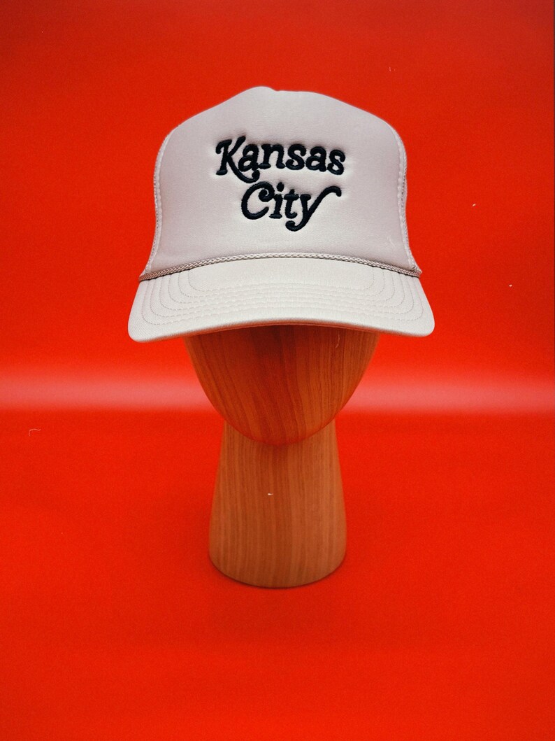 Pu&ograve; includere: Un cappello da camionista beige con un logo ricamato nero che dice "Kansas City".