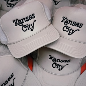 Pu&ograve; includere: Una pila di cinque cappellini da camionista grigi con pannelli posteriori in rete nera. Ogni cappellino presenta un logo ricamato nero che dice "Kansas City" in un font di scrittura retr&ograve;.