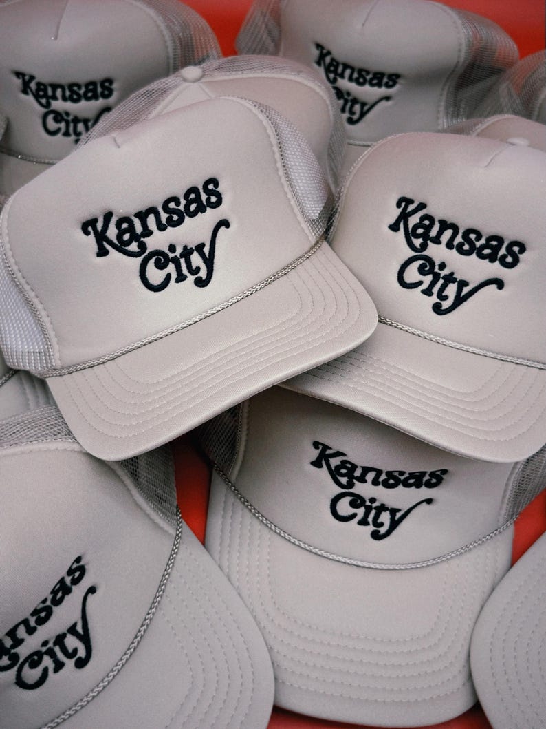 Pu&ograve; includere: Una pila di cinque cappellini da camionista grigi con pannelli posteriori in rete nera. Ogni cappellino presenta un logo ricamato nero che dice "Kansas City" in un font di scrittura retr&ograve;.