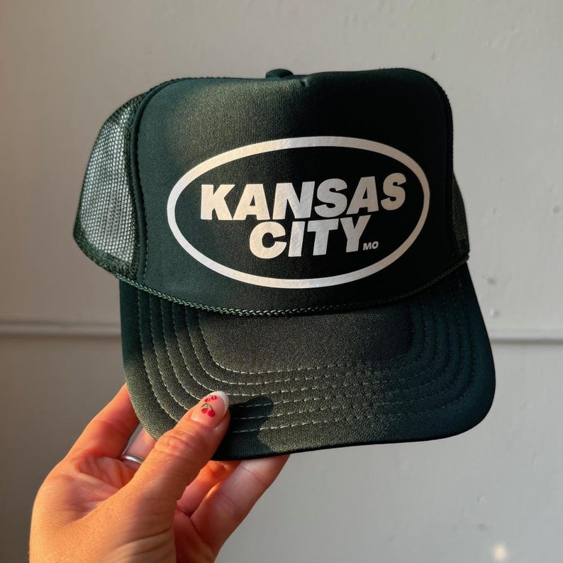 May include: A green trucker hat with a white oval patch that says "KANSAS CITY MO" in white block letters.