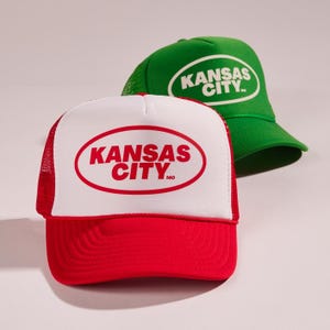 May include: Two trucker hats in a red and white colour scheme. Both hats feature a white oval with the text "KANSAS CITY" in red letters. The top hat is green and the bottom hat is red.