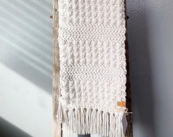 May include: A white crocheted scarf with a textured pattern and fringe. The scarf is hanging on a wooden ladder.