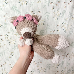 May include: A crocheted plush toy fox with a pink flower crown. The fox is brown and white with a brown nose and white feet. The fox is being held by a hand.