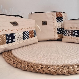 Peut inclure: Ensemble de trois sacs en tissu beige avec des accents patchwork aux motifs marine, marron et blancs. Les sacs sont posés sur un dessous-de-plat rond en osier. Le plus grand sac a une fermeture éclair et des détails brodés.