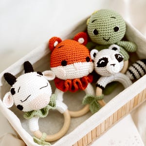 May include: A collection of handmade crochet baby rattles in a woven basket. The rattles feature animal designs: a white cow, an orange fox, a green turtle, and a black and white raccoon. Each rattle has a wooden ring and is crafted with soft yarn.