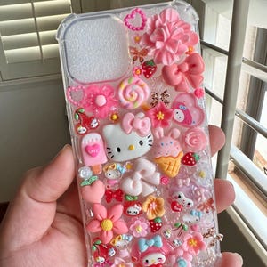 以下が含まれることがあります： ピンク、白、赤の3D装飾で飾られた透明な携帯電話ケース。ケースには、ハローキティのキャラクター、花、リボン、ハート、その他の可愛いデザインが施されています。