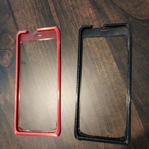 May include: Two plastic phone cases, one red and one black, are shown on a wooden surface. Both cases have a clear plastic window in the center.