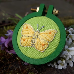 Op de afbeelding: Een geborduurde gele vlinder op een groene stoffen achtergrond in een houten borduurring.