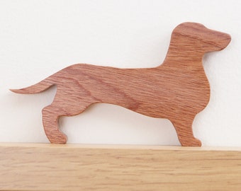 May include: A wooden dachshund silhouette, carved from light brown wood, with a smooth, polished finish. The dachshund is standing on a wooden shelf.