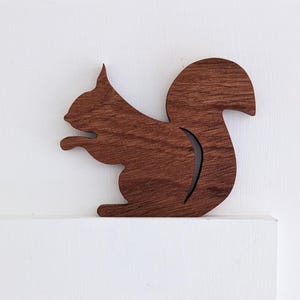May include: A wooden squirrel silhouette cutout in a dark brown colour. The squirrel is facing left and has its tail curled up behind it.