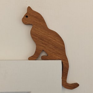 May include: A wooden cat silhouette in a brown colour, mounted on a white wall. The cat is sitting with its tail curled around its body.
