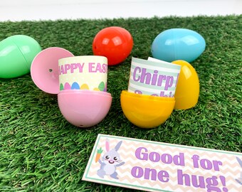 May include: A set of colorful plastic Easter eggs with different messages inside. The eggs are on a green grass surface. One egg is open and shows a yellow paper with the text "Chirp!" inside. Another egg is open and shows a pink paper with the text "Happy Easter" inside. A sign with the text "Good for one hug!" is in the foreground.