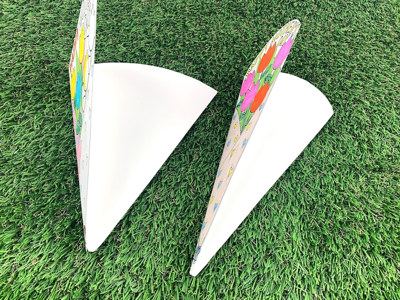 May include: Two white paper cones with colorful flower designs. The cones are folded and ready to be filled with treats or gifts.