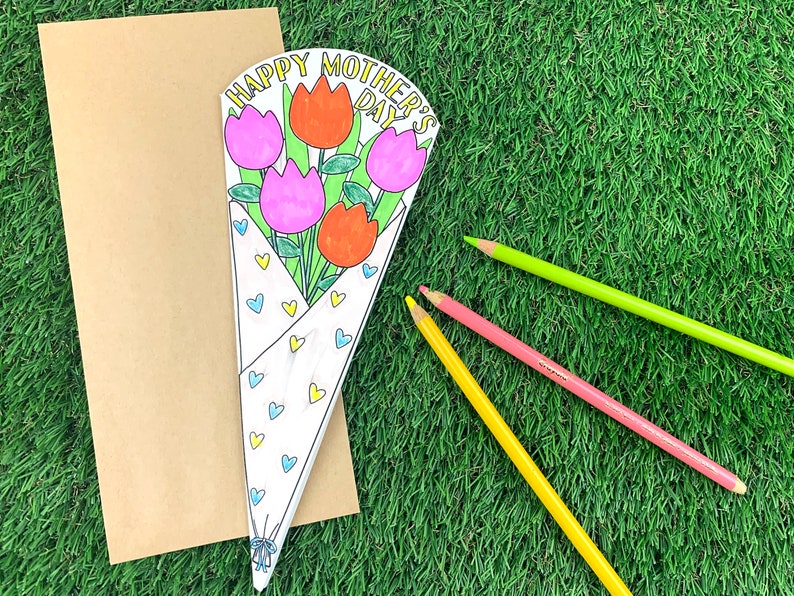 May include: A white card with a bouquet of pink and red tulips with green stems and leaves. The card says "Happy Mother's Day" in yellow letters. There are three colored pencils on a green grassy surface.