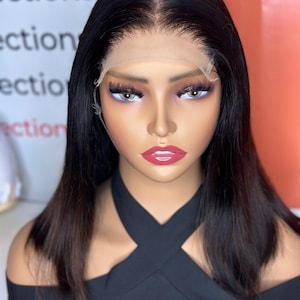 May include: A mannequin head with a black wig featuring a natural hairline and a lace front. The wig has a straight, shoulder-length style. The mannequin is wearing a black off-the-shoulder top and has full makeup with red lipstick.