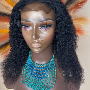 May include: A black curly human hair wig with a lace front. The wig is styled in loose curls and is worn on a mannequin head. The mannequin is wearing a turquoise beaded necklace.