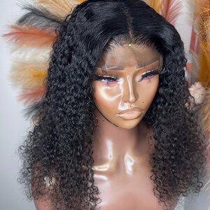 May include: A black curly wig with a lace front is displayed on a mannequin head. The wig features a natural-looking hairline and a deep part. The hair is styled in loose, bouncy curls. The mannequin is set against a backdrop of colorful decorative feathers.