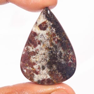 Op de afbeelding: Een druppelvormige edelsteen met een gepolijst oppervlak. De steen heeft een mix van kleuren, waaronder bordeauxrood, wit en donkerbruin, waardoor een uniek patroon ontstaat. De edelsteen wordt tussen twee vingers gehouden.