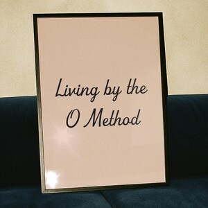 May include: A framed print with the text "Living by the O Method" on a pink background, set against a dark blue sofa. A clear champagne flute sits on a dark wooden table in the foreground.