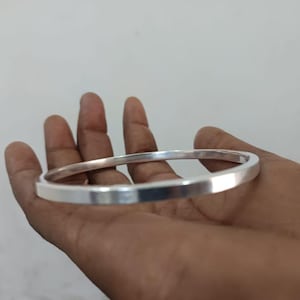Puede incluir: Un brazalete plateado con bordes cuadrados. El brazalete se sostiene en una mano, mostrando su superficie pulida y reflectante. Una pieza de joyería sencilla y elegante.