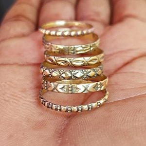 Puede incluir: Una pila de seis anillos dorados con varios patrones en relieve. Los anillos son delgados y presentan diseños como corazones, puntos y formas geométricas. Los anillos están apilados uno encima del otro.