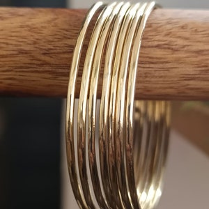 May include: A set of ten gold-toned bangle bracelets stacked on a wooden surface. The bracelets are thin and have a smooth, polished finish.