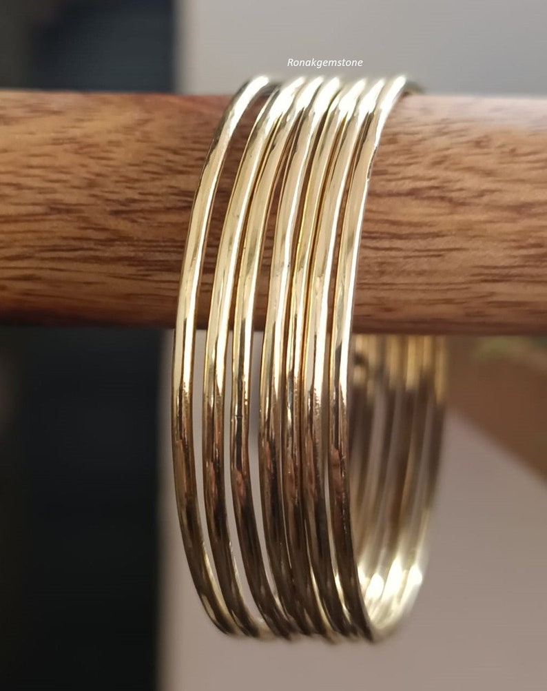 May include: A set of ten gold-toned bangle bracelets stacked on a wooden surface. The bracelets are thin and have a smooth, polished finish.
