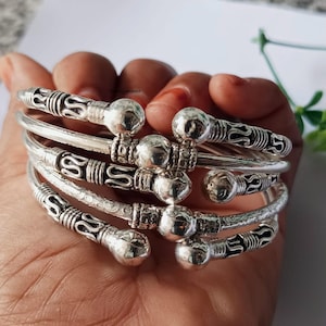 Può includere: Una collezione di braccialetti color argento. Ogni braccialetto presenta dettagli decorativi con una sfera centrale ed estremità ornate. I braccialetti sono impilati, mostrando il loro design e la loro consistenza. L'estetica generale è quella di gioielli fatti a mano.