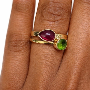 Peut inclure: Deux bagues en or avec des pierres précieuses. L'une des bagues présente une pierre précieuse rouge foncé en forme de poire. L'autre bague a une pierre précieuse verte claire ronde. Les bagues sont empilées sur un doigt.