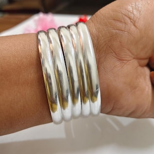 Puede incluir: Un conjunto de cinco pulseras de color plateado. Las pulseras son lisas y redondeadas, reflejando la luz. Las joyas se llevan en la muñeca. Cada pulsera tiene un diámetro de aproximadamente 6 cm.