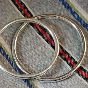 Puede incluir: Se muestran dos brazaletes plateados, de forma circular. Los brazaletes se superponen sobre una tela gris con rayas rojas, negras y blancas. La tela tiene un sutil estampado de rayas. El texto "Ronakgemstone" es visible.