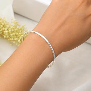May include: A simple, polished silver-tone bangle bracelet with a smooth, rounded surface. The bracelet features a continuous, circular design that rests comfortably around the wrist, showcasing a clean, reflective metallic finish throughout.
