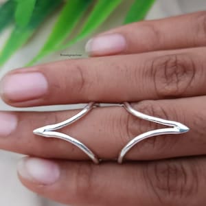 May include: A silver-colored ring with a unique design, featuring a geometric shape that wraps around the finger. The ring is shown on a hand, with the focus on the jewelry's intricate details and polished finish.