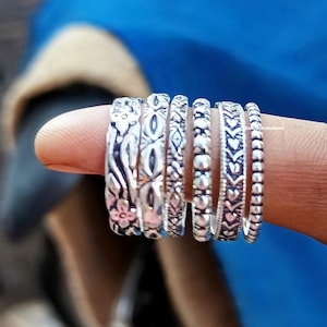 May include: A set of six silver stacking rings with different intricate designs. The rings are worn on a finger and feature floral, geometric, and heart patterns.