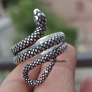 May include: A silver-colored snake ring with a detailed scale pattern. The snake's head is raised, and the body coils around the finger. The ring is made of a shiny metal and has a textured surface.