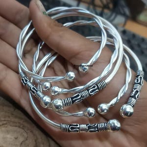 Puede incluir: Conjunto de cinco pulseras de plata con diseños trenzados y pequeñas cuentas redondas. Las pulseras están apiladas unas sobre otras.
