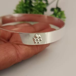Pode incluir: Uma pulseira de bracelete prateada com uma faixa plana e larga. Um pequeno design floral em relevo é centrado na pulseira. A pulseira é segurada em uma mão, com uma planta verde desfocada ao fundo.