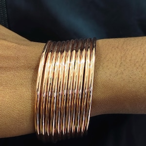 Puede incluir: Una pila de brazaletes de cobre, que reflejan la luz con un brillo metálico cálido. Los brazaletes son cilíndricos y lisos, creando un efecto en capas en la muñeca. El color cobre es prominente, con sutiles variaciones de tono.