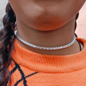 May include: A silver choker necklace with a hammered texture. The necklace is worn on a person wearing an orange sweater with a black geometric pattern. The choker is a simple, elegant piece of jewelry.