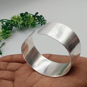 Puede incluir: Un brazalete ancho y abierto de plata con un acabado de metal cepillado. El brazalete se sostiene en una mano, con plantas decorativas verdes en el fondo. El brazalete mide aproximadamente 5 cm de ancho.