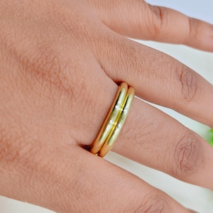 Puede incluir: Dos anillos dorados en un dedo. Los anillos tienen un diseño sencillo y clásico, hechos de metal amarillo brillante. Los anillos están juntos. La imagen es un primer plano de los anillos en una mano. Los anillos son probablemente de metal precioso.