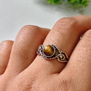 Puede incluir: Un anillo de plata con una piedra preciosa de ojo de tigre pulida. El anillo tiene un diseño de filigrana ornamentado con detalles decorativos alrededor de la piedra y a lo largo de la banda. El ojo de tigre muestra bandas doradas y negras.