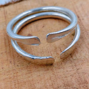 May include: A silver-colored, open-ended ring with a double-band design. The ring has a smooth, polished finish and is set against a wooden surface. The ring's design allows for slight size adjustments.