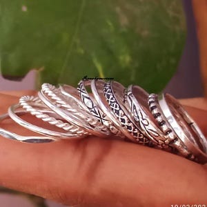 Puede incluir: Un conjunto de nueve anillos de plata con diferentes diseños. Los anillos están apilados uno encima del otro y están siendo sostenidos por una mano.