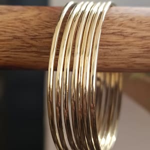 May include: A set of ten gold-toned bangle bracelets stacked on a wooden surface. The bracelets are thin and have a smooth, polished finish.